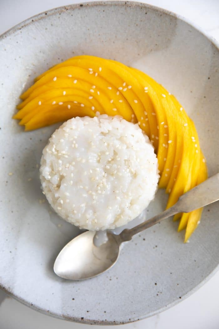 Small serving of mango sticky rice topped with coconut cream and sesame seeds and served with sliced mango.