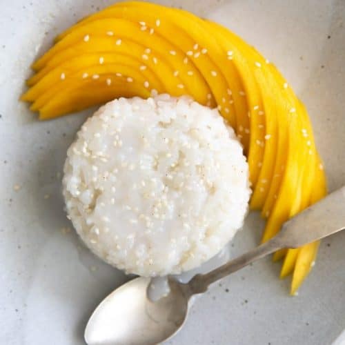 Small serving of mango sticky rice topped with coconut cream and sesame seeds and served with sliced mango.
