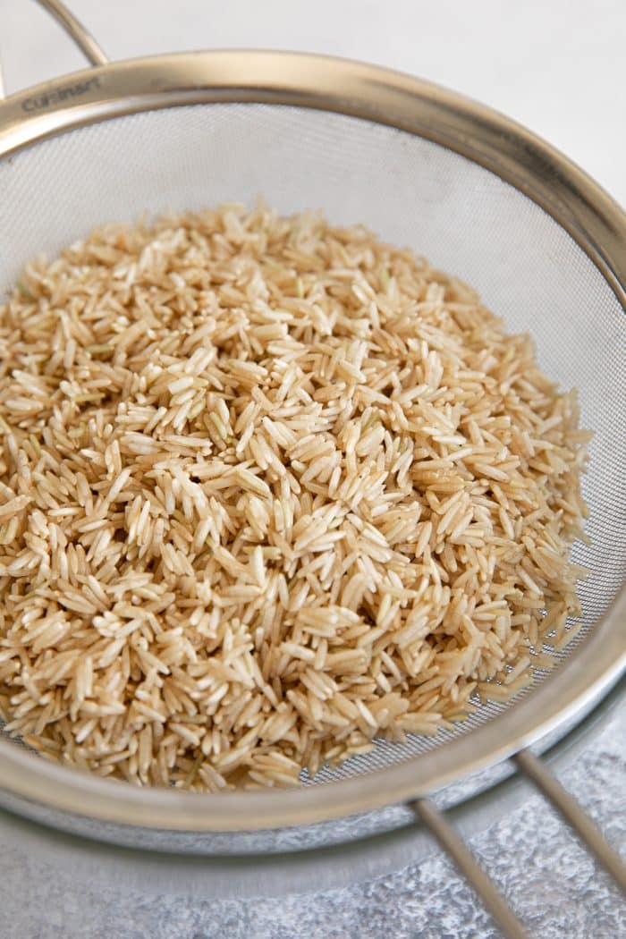 Uncooked brown basmati rice draining over a glass bowl in a fine-mash strainer.
