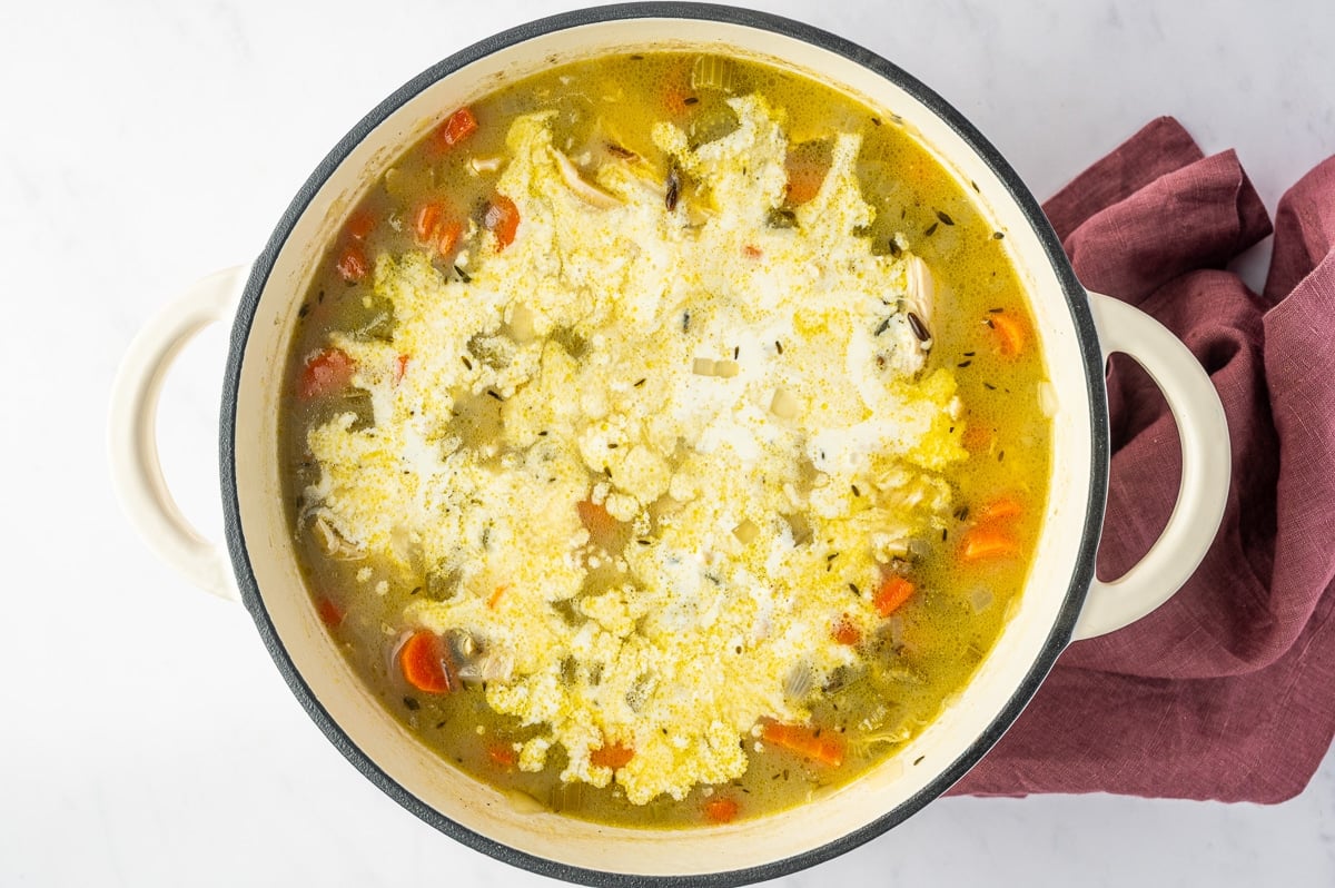 Heavy cream freshly poured across chicken and wild rice soup in a white Dutch oven, creating creamy white swirls among the shredded chicken, carrots, celery, wild rice, and broth.