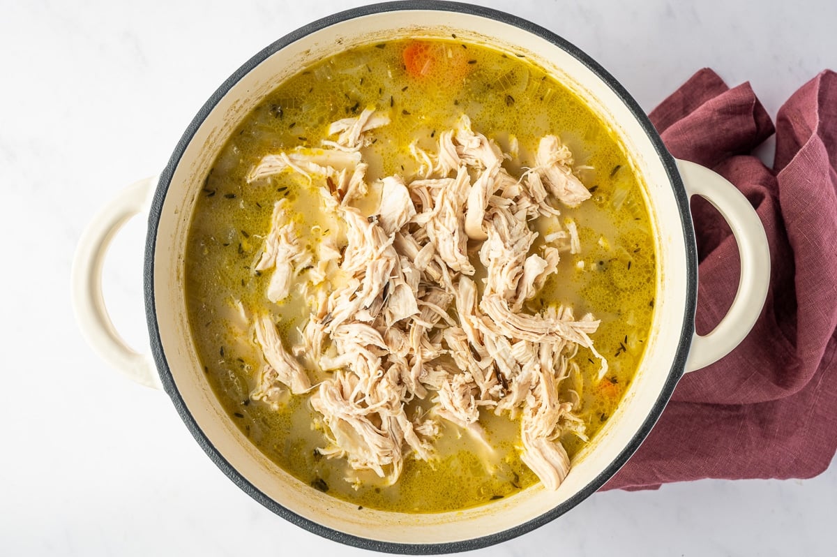 Freshly shredded chicken piled into the center of a Dutch oven filled with cooked wild rice, vegetables, herbs, and golden chicken broth.