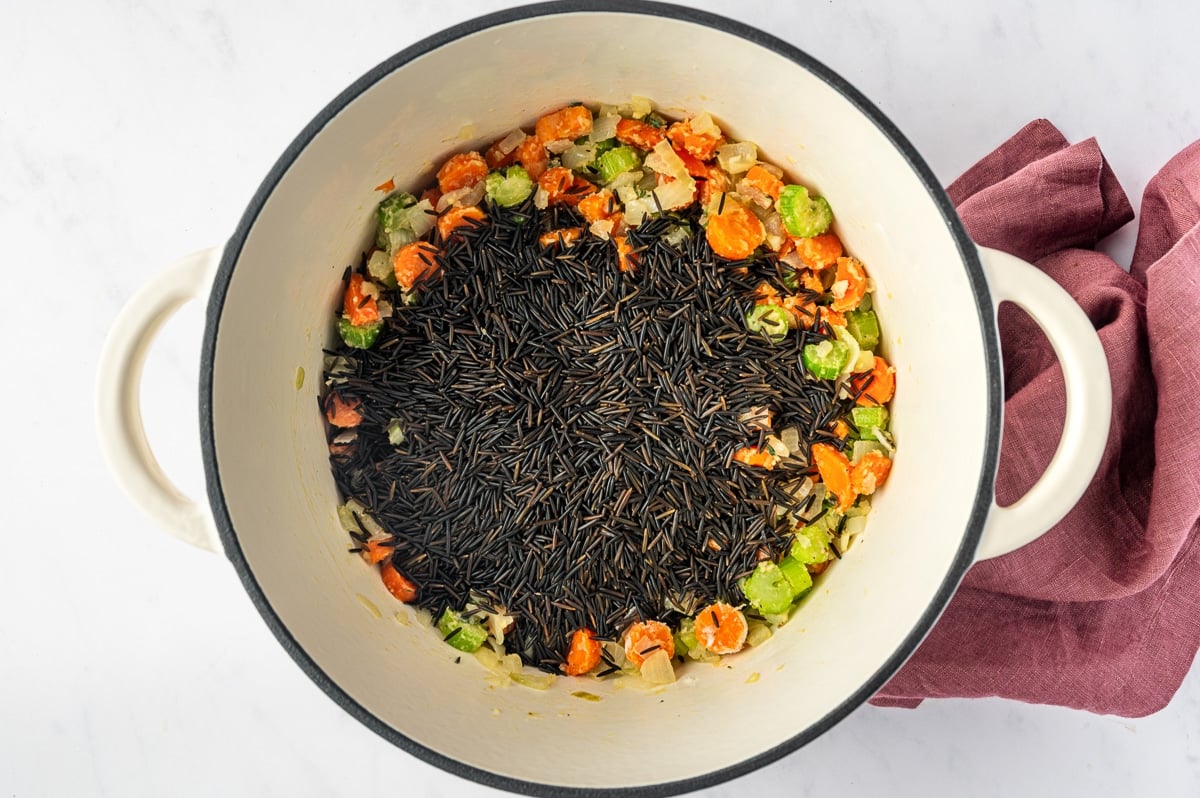 Dark uncooked wild rice added to a Dutch oven filled with softened carrots, celery, onion, herbs, and the flour mixture for chicken and wild rice soup.