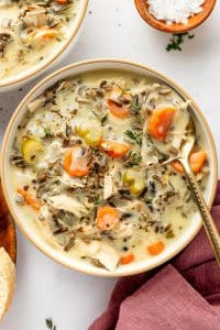 A large cream-colored bowl filled with chicken and wild rice soup, including shredded chicken, dark wild rice grains, sliced carrots, celery, onion, fresh thyme, and black pepper in a creamy broth, with a spoon resting inside the bowl.