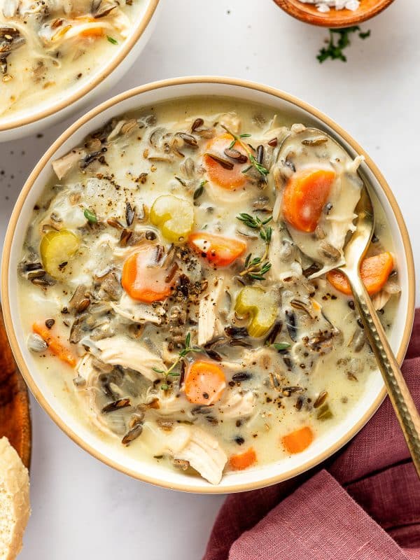 A large cream-colored bowl filled with chicken and wild rice soup, including shredded chicken, dark wild rice grains, sliced carrots, celery, onion, fresh thyme, and black pepper in a creamy broth, with a spoon resting inside the bowl.