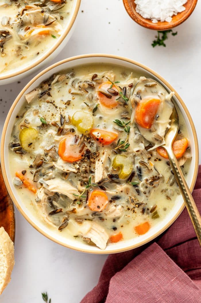 A large cream-colored bowl filled with chicken and wild rice soup, including shredded chicken, dark wild rice grains, sliced carrots, celery, onion, fresh thyme, and black pepper in a creamy broth, with a spoon resting inside the bowl.