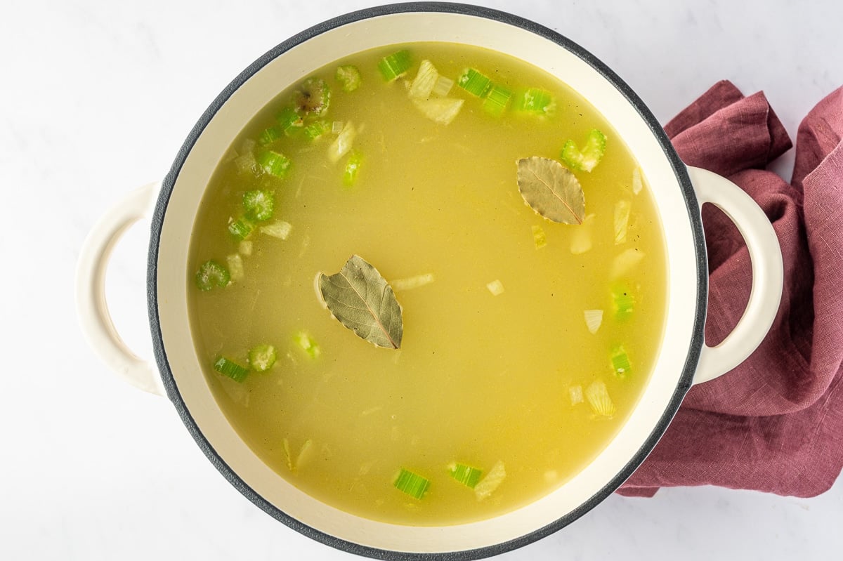 Golden chicken broth filling a white Dutch oven with pieces of onion and celery visible beneath the surface and two whole bay leaves floating on top.