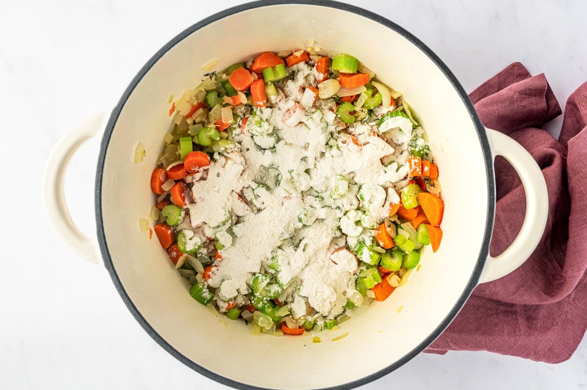 Softened carrots, celery, and onion in a white Dutch oven topped with all-purpose flour, ready to be stirred together to form the light roux that thickens the soup.