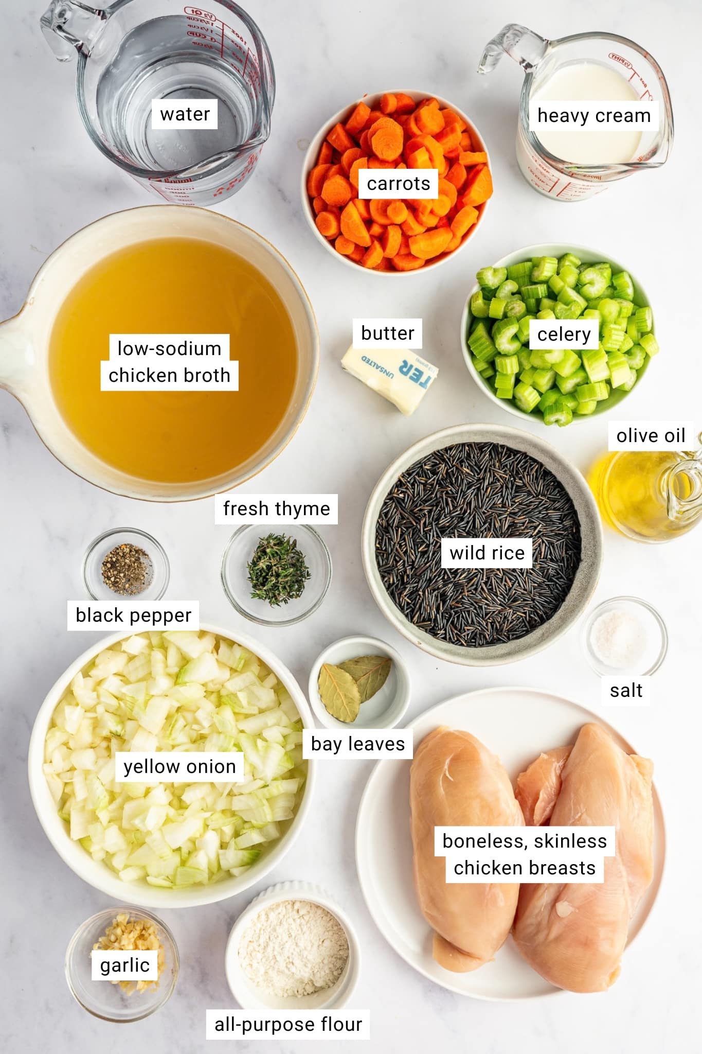 Ingredients for chicken and wild rice soup arranged together and individually labeled, including boneless skinless chicken breasts, wild rice, yellow onion, carrots, celery, garlic, fresh thyme, bay leaves, all-purpose flour, butter, olive oil, low-sodium chicken broth, water, heavy cream, salt, and black pepper.