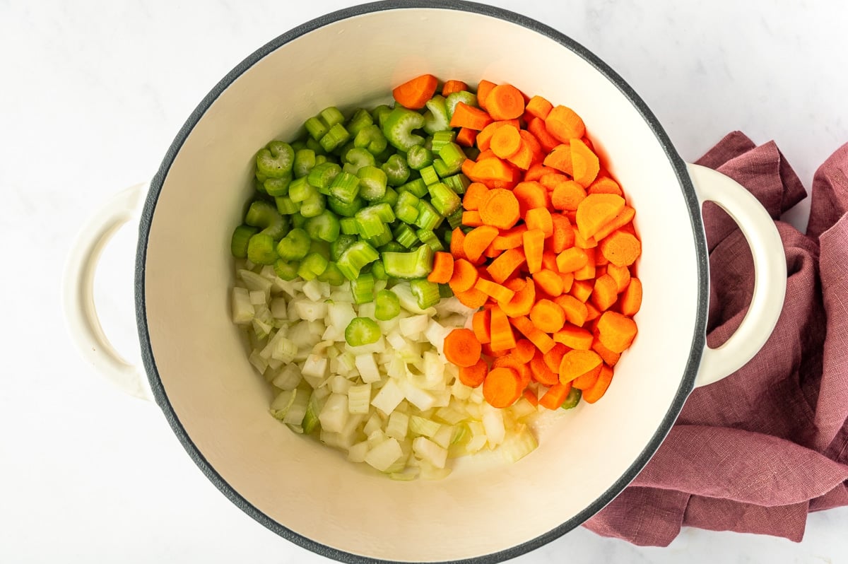 Chopped yellow onion, sliced carrots, and sliced celery arranged in a large white enameled Dutch oven before being sauteed for chicken and wild rice soup.