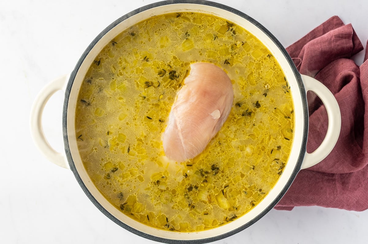 A boneless skinless chicken breast gently simmering in the seasoned broth with softened vegetables, thyme, and wild rice as the chicken cooks directly in the soup.