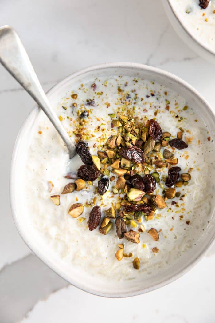 Small white dessert bowl filled with cardamom spiced Indian rice pudding and garnished with crushed pistachios and raisins.