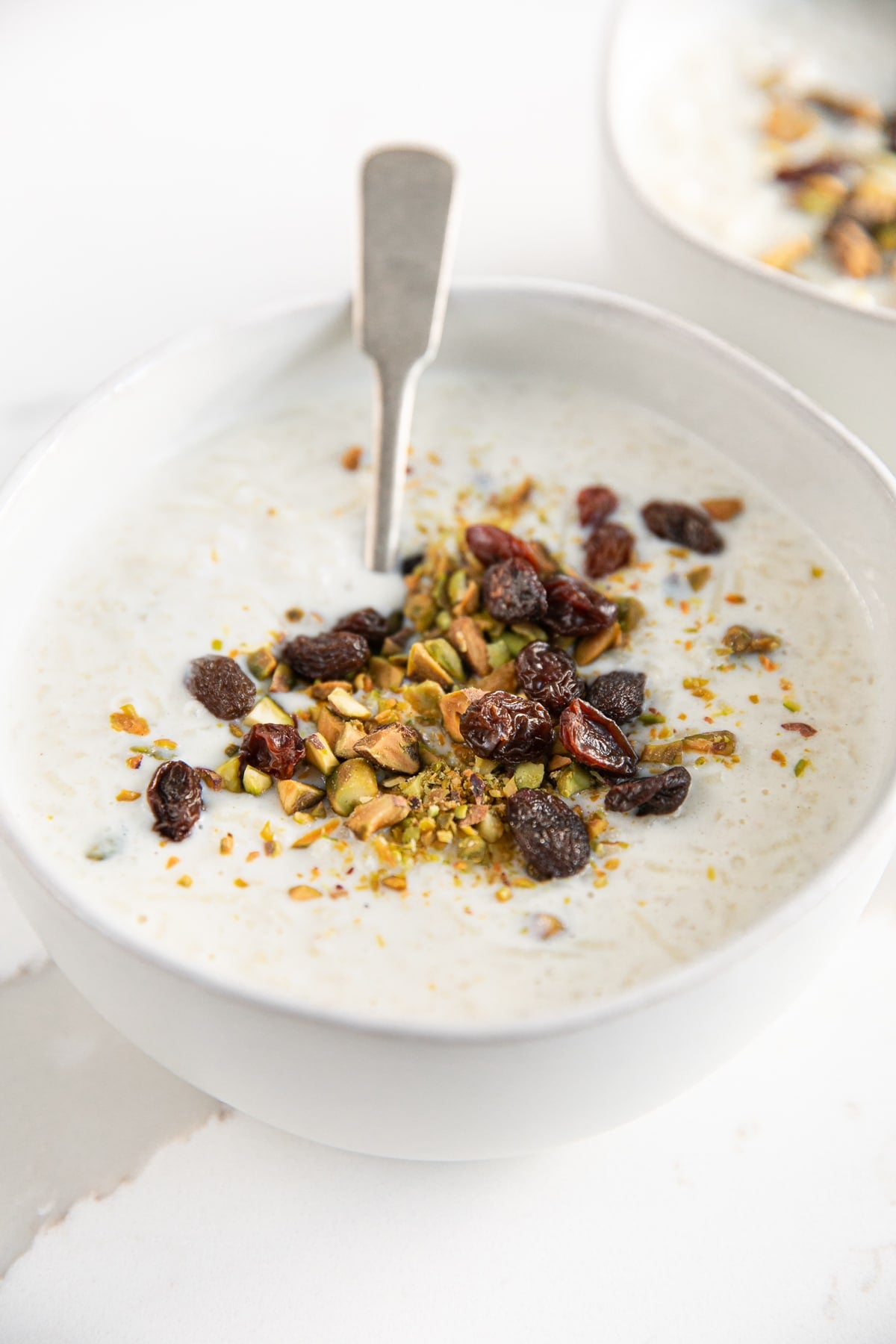 Small white dessert bowl filled with cardamom spiced Indian rice pudding and garnished with crushed pistachios and raisins.