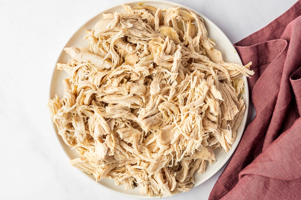 A white plate piled high with cooked chicken breast shredded into a mixture of thin strands and larger tender pieces, ready to be returned to the soup.