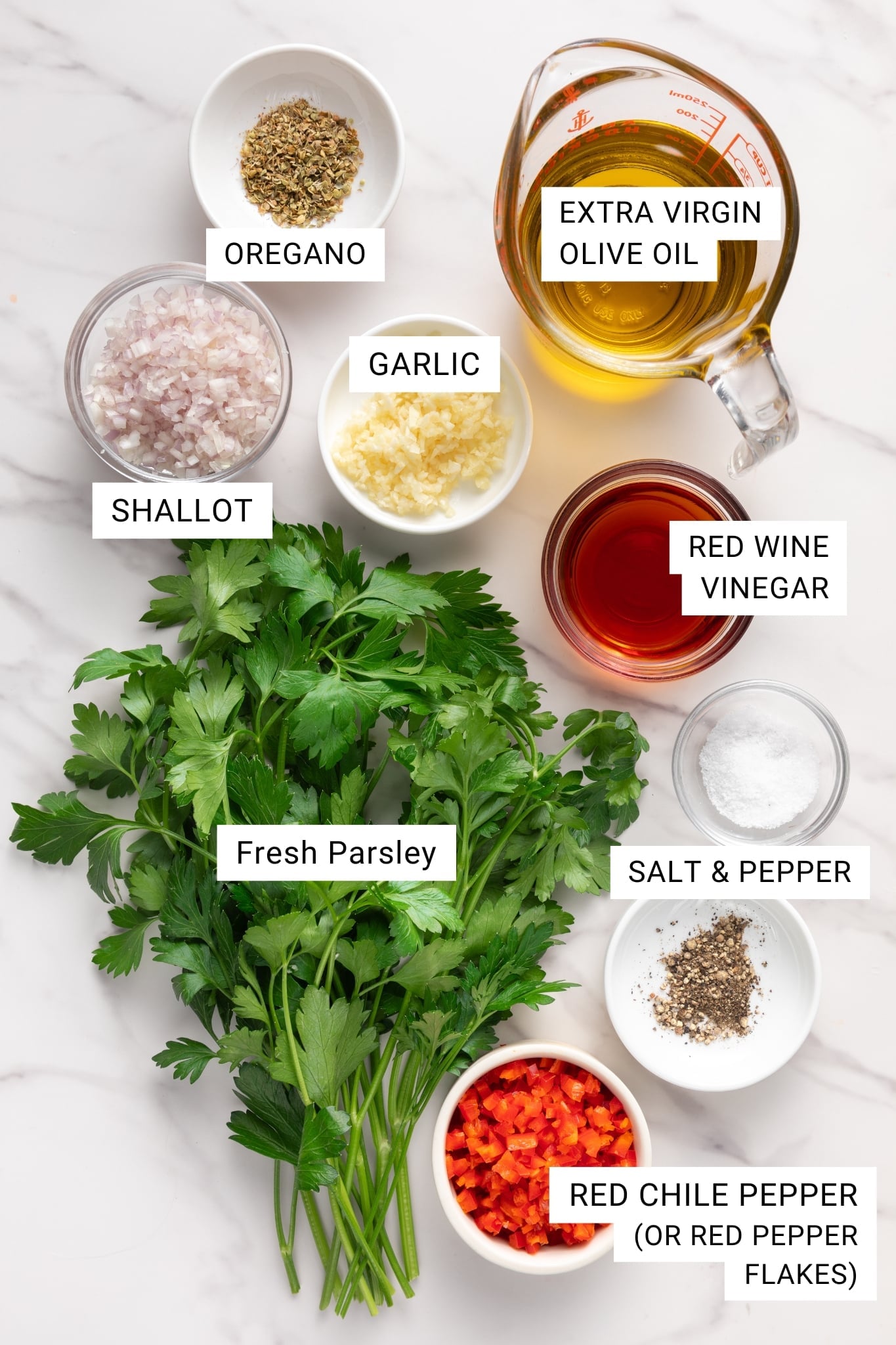 Overhead photo on a white marble surface showing chimichurri ingredients with white text labels: fresh parsley bunch, minced garlic in a small white bowl, diced shallot in a small glass bowl, dried oregano in a white ramekin, extra virgin olive oil in a glass measuring cup, red wine vinegar in a clear glass, salt in a small glass bowl, cracked black pepper in a white dish, and chopped red chile pepper in a small white bowl labeled &ldquo;RED CHILE PEPPER (OR RED PEPPER FLAKES)&rdquo;.