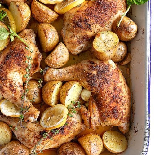 Large baking pan filled with golden crispy whole chicken legs slow roasted with baby potatoes, whole heads of garlic, and fresh lemon slices.