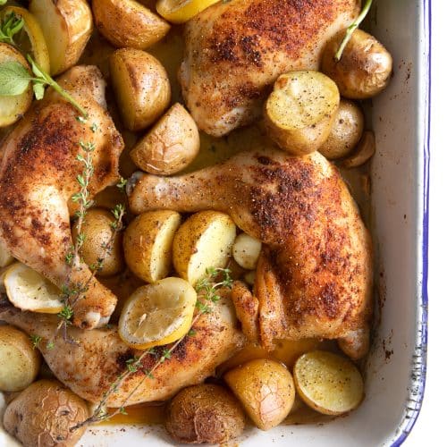 Baking pan filled with whole chicken legs, tender potatoes, sliced lemon, and fresh herbs.
