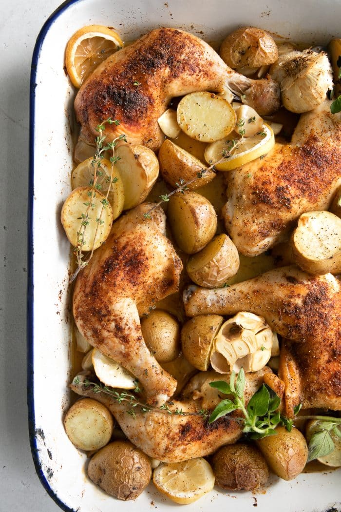 Baking pan filled with whole chicken legs, tender potatoes, sliced lemon, garlic cloves, and fresh herbs.