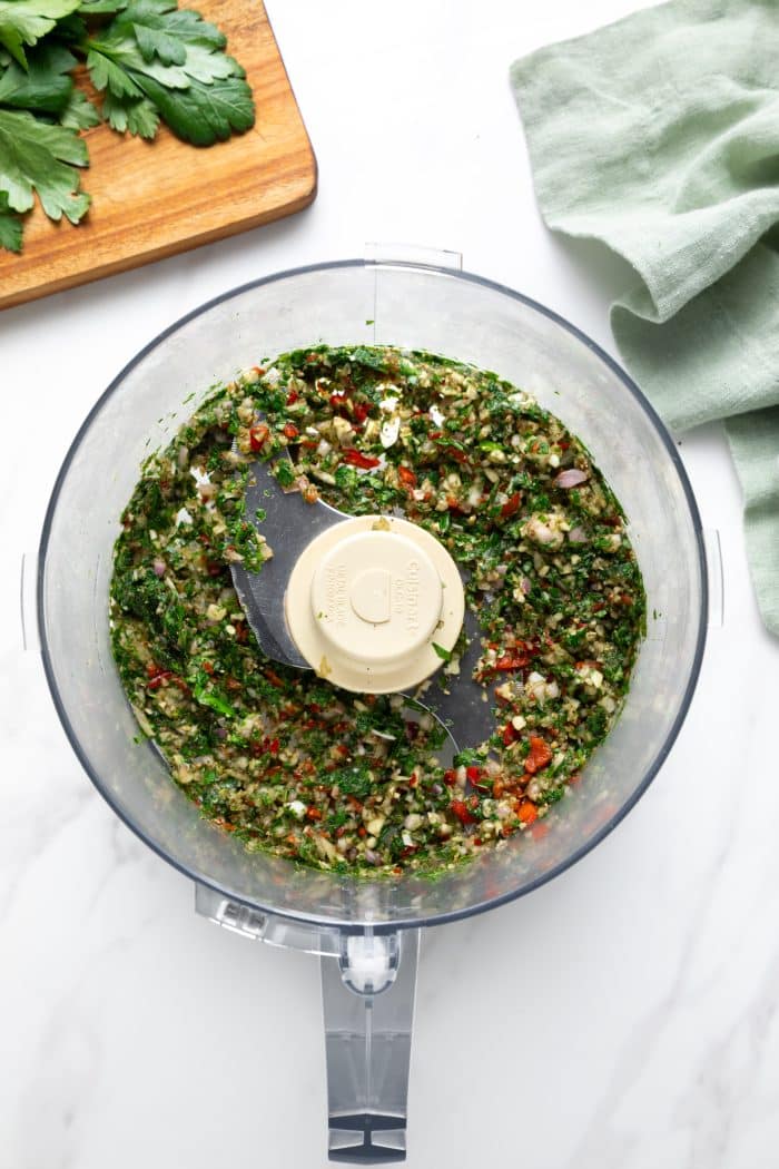 Overhead view of a food processor bowl after pulsing chimichurri; the mixture is chopped and chunky with visible parsley, garlic, shallot, and red chile pieces around the blade, with a wooden cutting board and parsley at the top left and a pale green linen cloth at the top right.