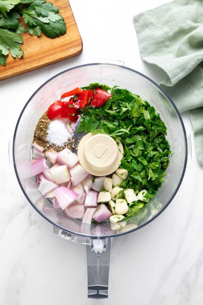 Overhead shot of a food processor bowl filled with chimichurri ingredients before blending: chopped parsley, whole garlic cloves, chunks of shallot, dried oregano, salt, black pepper, and pieces of red chile pepper; a wooden cutting board with parsley is in the top left and a pale green linen cloth is in the top right.