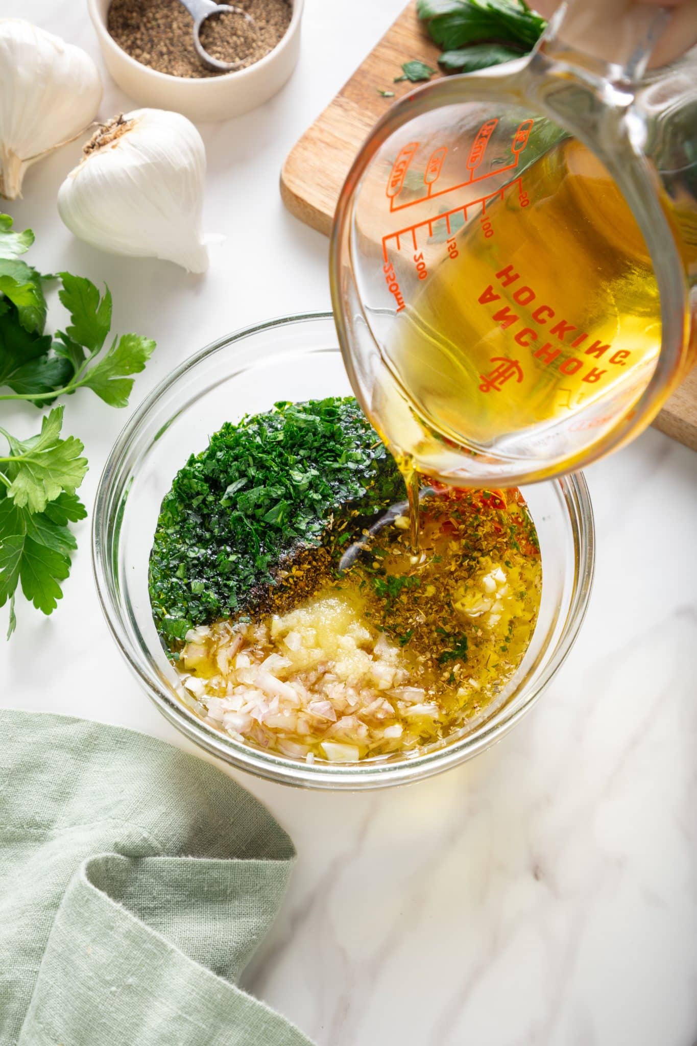 Olive oil being poured from a glass measuring cup with red markings into a clear glass bowl filled with chopped parsley, minced garlic, diced shallot, dried oregano, red wine vinegar, and black pepper; garlic bulbs, a small bowl of pepper, and fresh parsley sit in the background with a wooden cutting board and a pale green linen cloth.