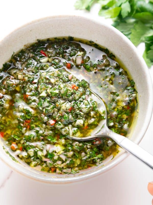 Close-up of chunky green chimichurri sauce in a light beige bowl with a silver spoon scooping up the sauce; visible bits of parsley, garlic, shallot, and red chile pieces float in olive oil, with fresh parsley leaves blurred in the background.
