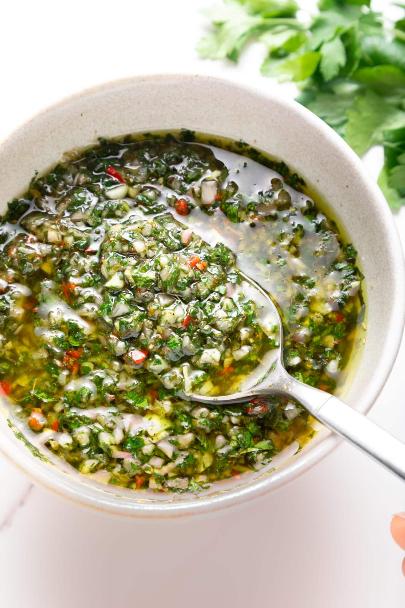 Close-up of chunky green chimichurri sauce in a light beige bowl with a silver spoon scooping up the sauce; visible bits of parsley, garlic, shallot, and red chile pieces float in olive oil, with fresh parsley leaves blurred in the background.