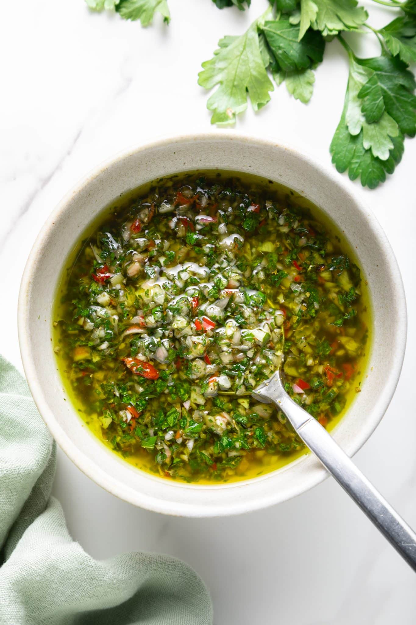 Overhead photo of a light beige bowl filled with chimichurri sauce and olive oil, with a silver spoon resting inside; chopped parsley, garlic, shallot, and red chile pieces are visible, with parsley sprigs at the top and a pale green cloth at the lower left.