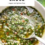 Vertical Pinterest pin with a white text box at the top and a close-up food photo underneath. The photo shows chunky green chimichurri sauce in a light beige bowl with a silver spoon scooping the sauce; visible parsley, garlic, shallot pieces, and red chile bits are suspended in olive oil, with blurred parsley leaves in the background. Text overlays read “5-minute,” large “CHIMICHURRI SAUCE,” and “vegan and gluten-free,” with “the forked spoon” at the bottom.