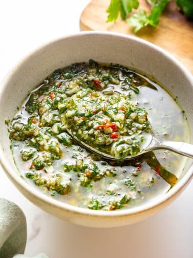 A silver spoon holds a spoonful of chimichurri above a light beige bowl; the sauce looks chunky with visible parsley, garlic, shallot, and red chile pieces suspended in olive oil, with a wooden cutting board and parsley in the background and a pale green cloth on the left.