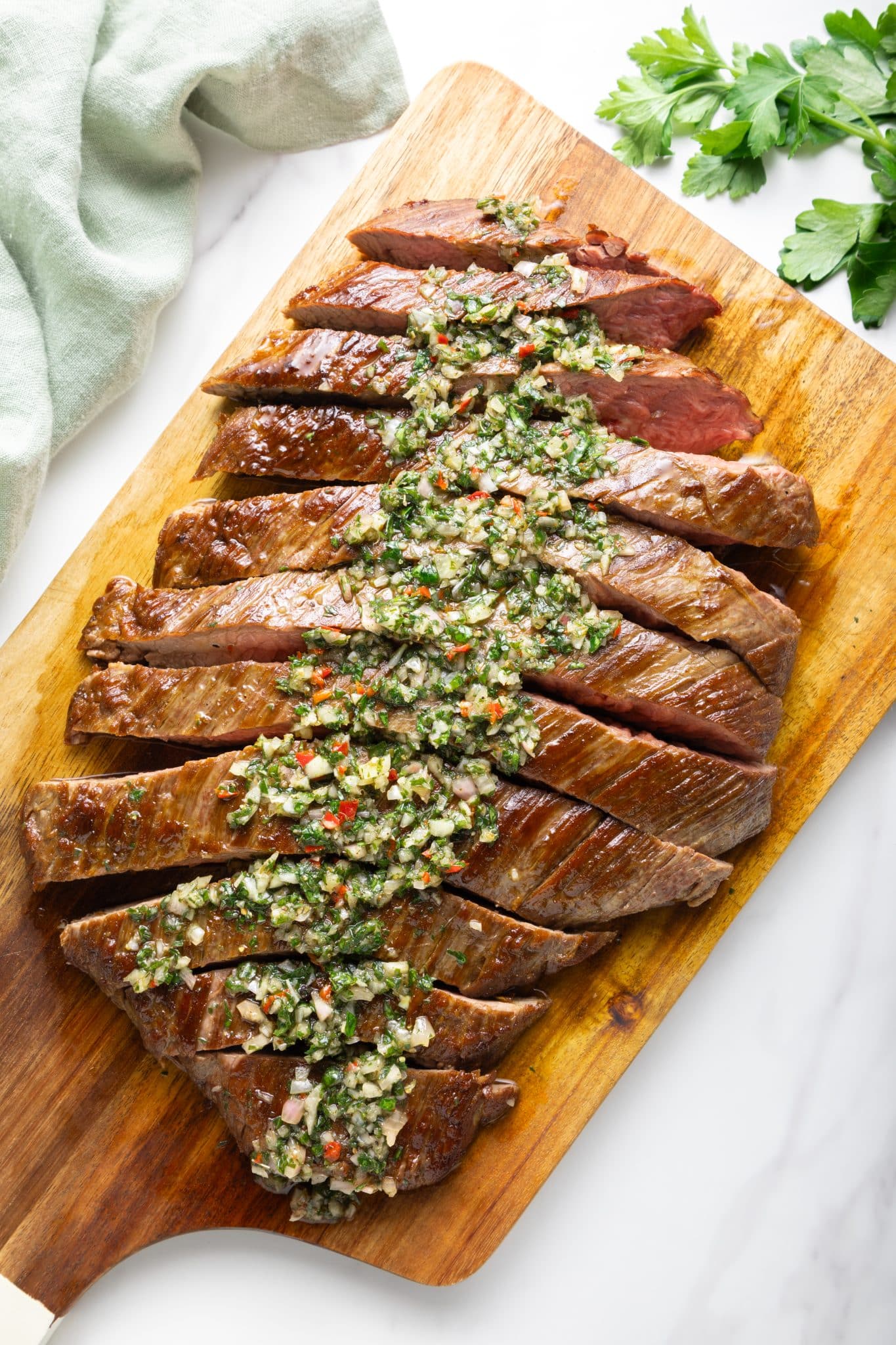 Sliced grilled steak arranged on a wooden cutting board and topped down the center with chunky chimichurri sauce; fresh parsley sprigs sit at the top right and a pale green linen cloth is draped at the top left on a white marble background.