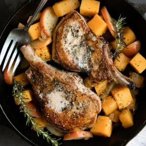 A large cast iron pan filled with two bone-in pork chops cooked to golden brown perfection and topped with melted butter and minced rosemary and surrounded by cook cubes of butternut squash and sliced softened apples.