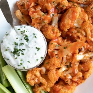 Cauliflower florets smothered and baked in buffalo hot sauce and served on a white platter with a small glass jar filled with ranch dressing and a side of celery sticks.