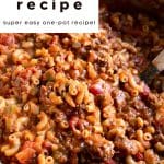 Close-up of a large pot filled with old-fashioned American goulash made with elbow macaroni, ground beef, diced tomatoes, and a rich tomato sauce. A wooden spoon is partially visible stirring the saucy pasta. Overlay text reads: โOld-Fashioned American Goulash Recipe โ Super Easy One-Pot Recipe!โ with โthe forked spoonโ branding at the bottom.