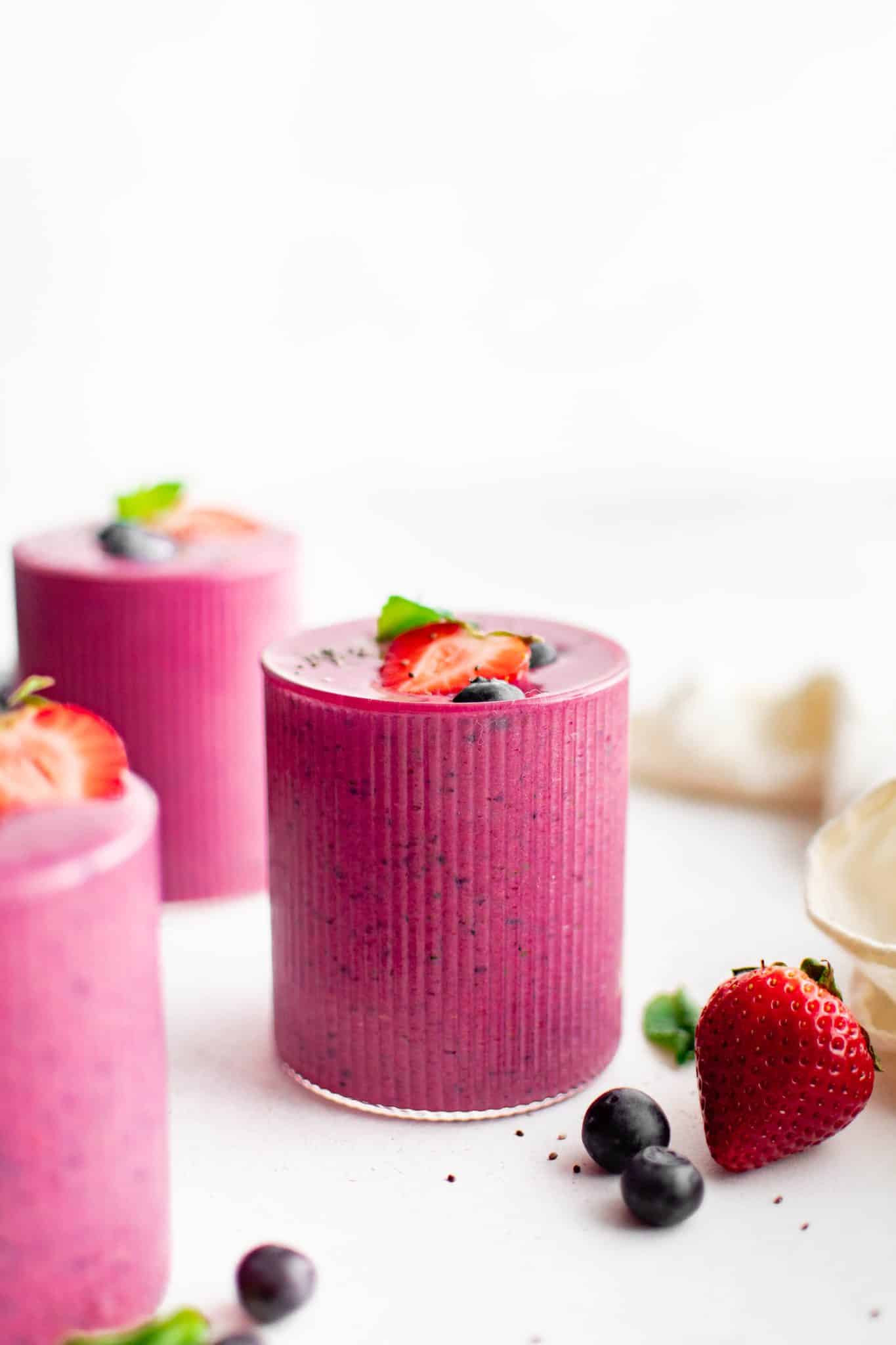 A thick and creamy pink beet smoothie in a textured glass topped with sliced strawberries, blueberries, and chia seeds, and set against a white background with scattered fruit for styling.