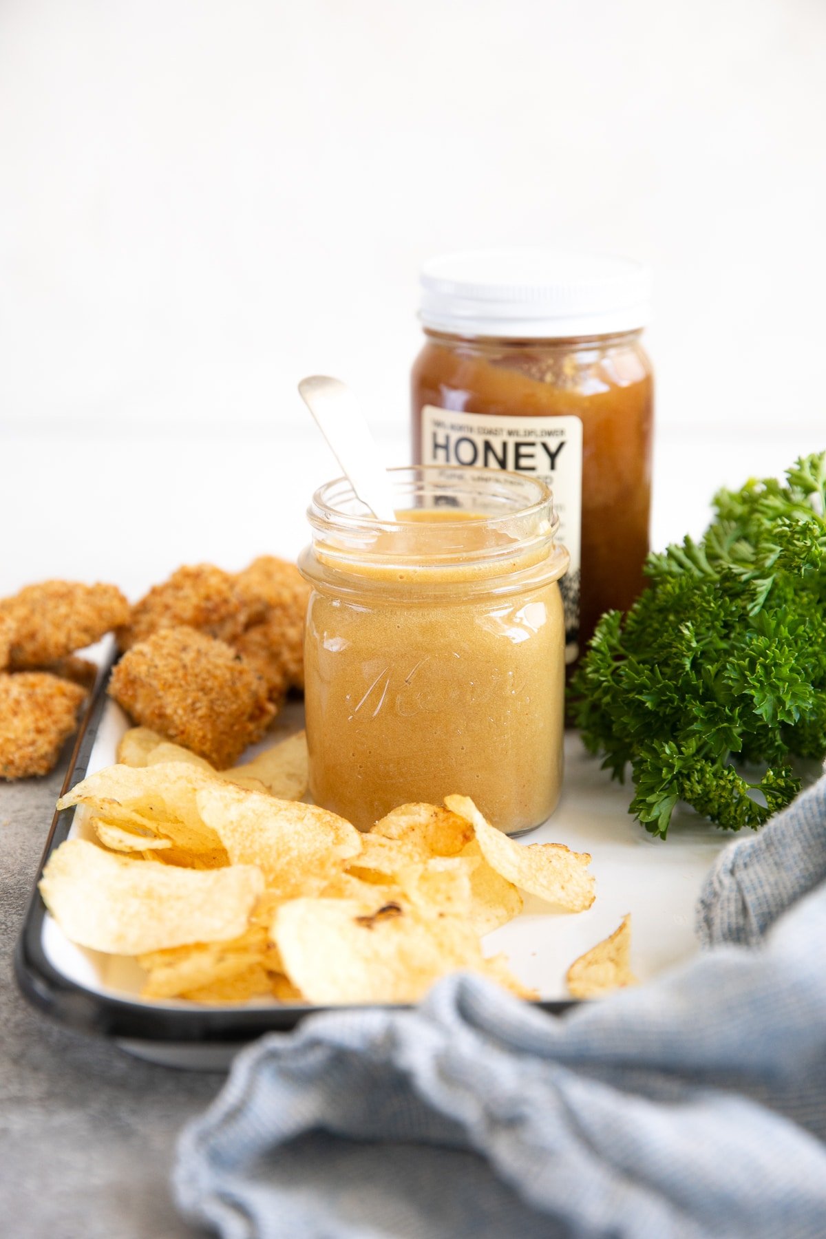 Honey mustard sauce on a white tray with potato chips, chicken nuggets, and a jar of honey in the background.