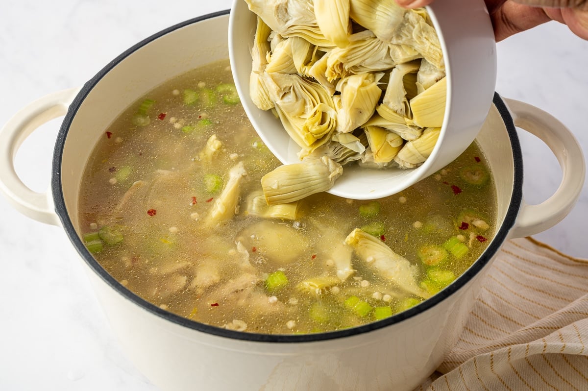 Quartered artichoke hearts being poured from a white bowl into lemon chicken soup with pearl couscous, shredded chicken, vegetables, and homemade broth.