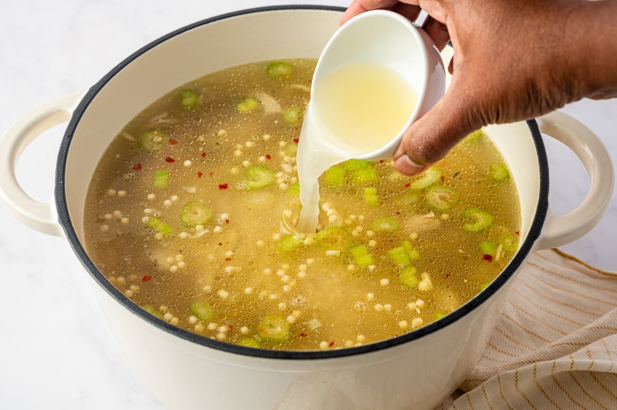 Fresh lemon juice being poured from a small white cup into a pot of chicken soup with pearl couscous, shredded chicken, carrots, celery, and red pepper flakes.