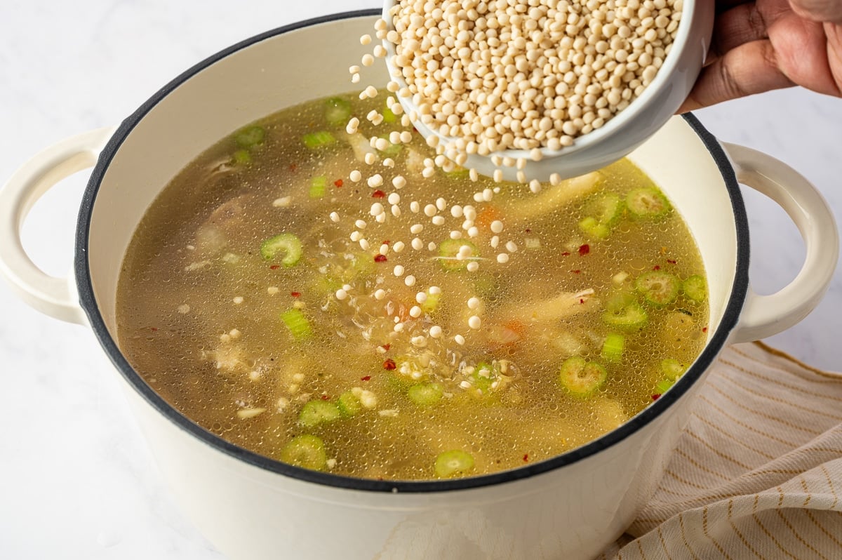 Pearl couscous being poured from a white bowl into a Dutch oven filled with homemade chicken broth, shredded chicken, carrots, celery, onion, and seasonings.