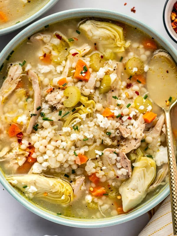 Bowl of lemon chicken soup filled with shredded chicken, pearl couscous, carrots, celery, and large artichoke heart quarters in a light golden broth, topped with crumbled feta, chopped chives, black pepper, and red pepper flakes.