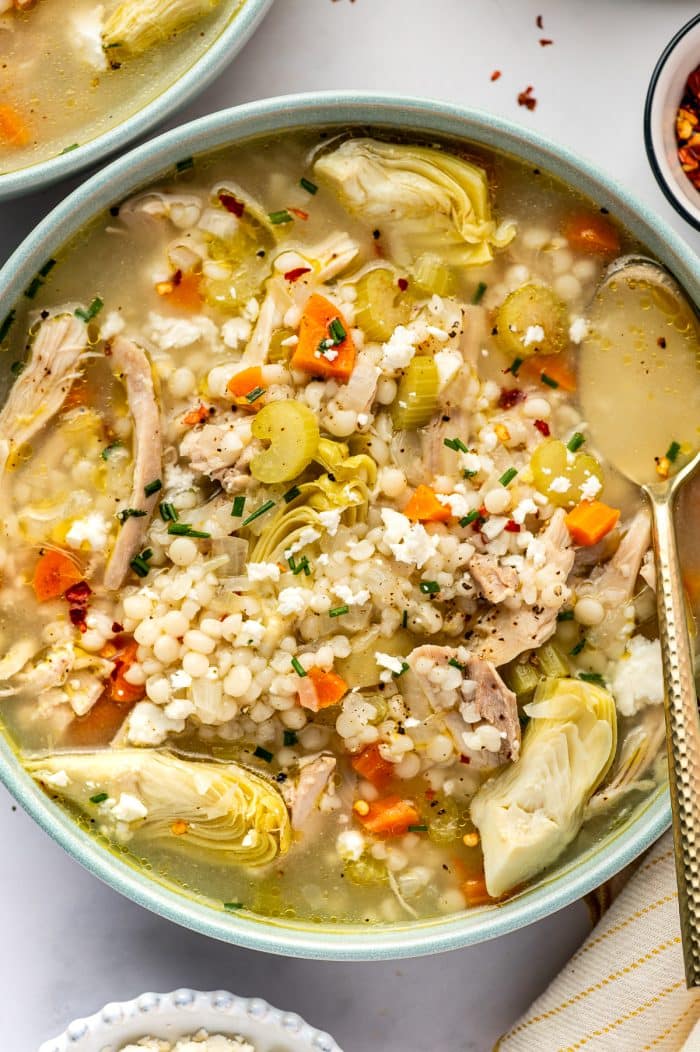 Bowl of lemon chicken soup filled with shredded chicken, pearl couscous, carrots, celery, and large artichoke heart quarters in a light golden broth, topped with crumbled feta, chopped chives, black pepper, and red pepper flakes.