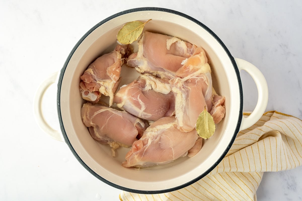 Bone-in, skinless chicken thighs submerged in water with two bay leaves in a white Dutch oven, ready to simmer and make the homemade chicken broth for lemon chicken soup.