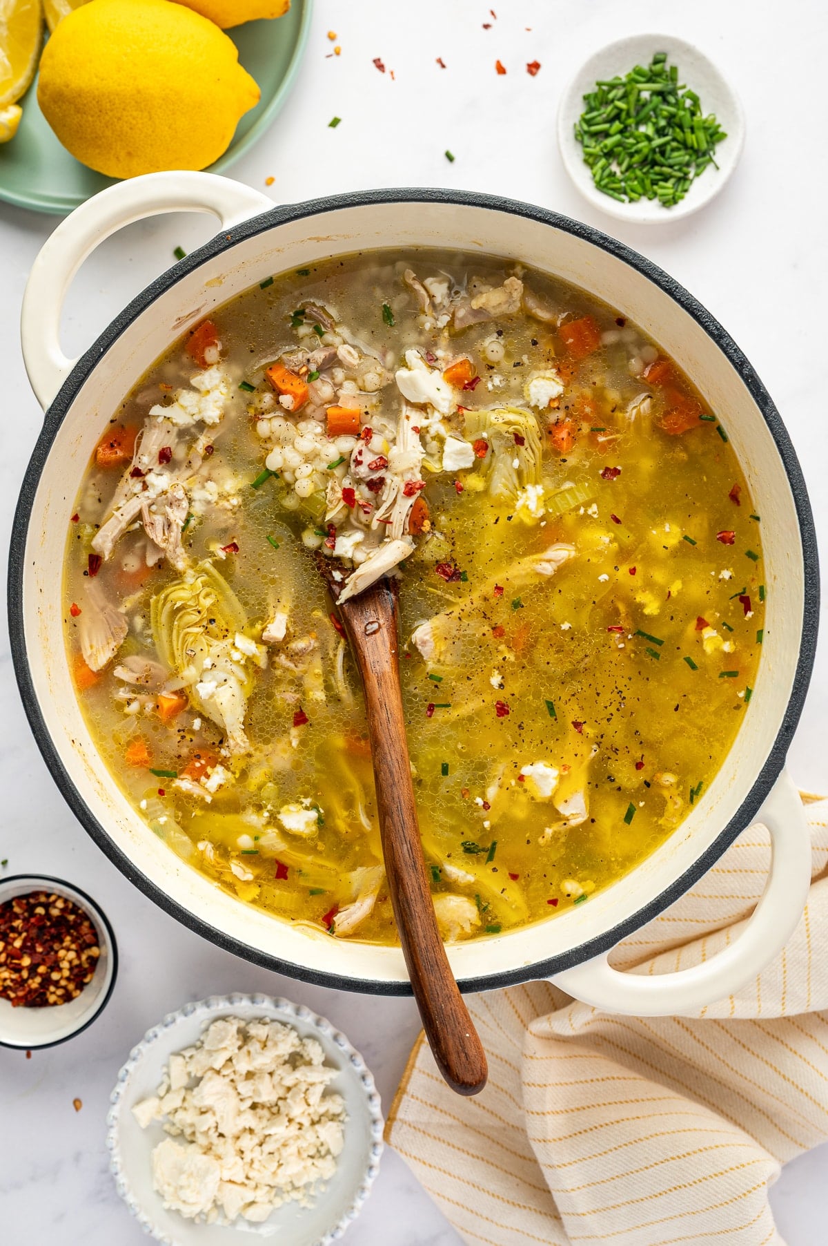 Finished lemon chicken soup in a white Dutch oven with shredded chicken, pearl couscous, carrots, celery, artichoke hearts, feta cheese, chopped chives, black pepper, and red pepper flakes, with a wooden spoon resting in the pot.