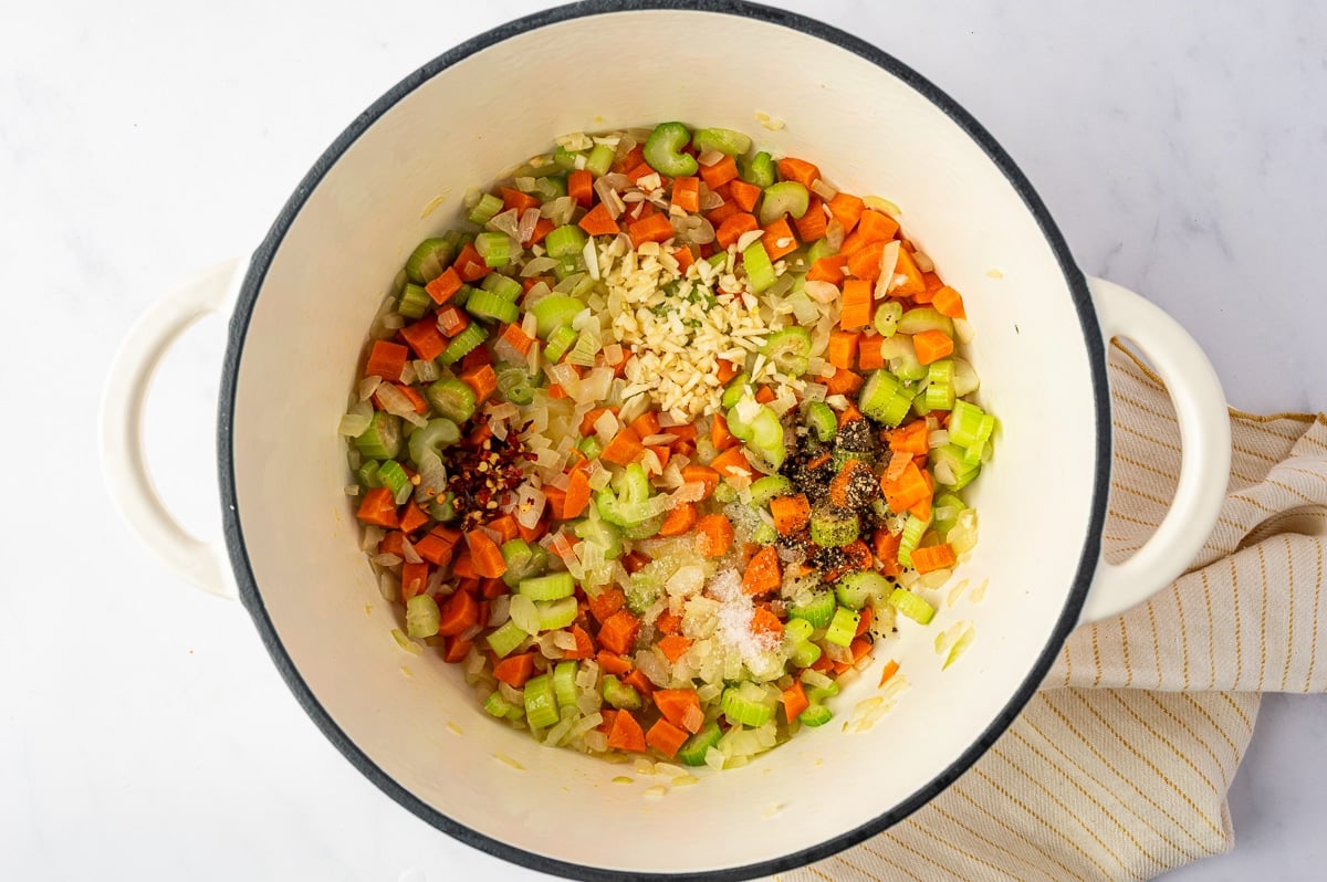 Sauteed carrots, celery, and onion topped with minced garlic, salt, black pepper, and crushed red pepper flakes in a white Dutch oven.