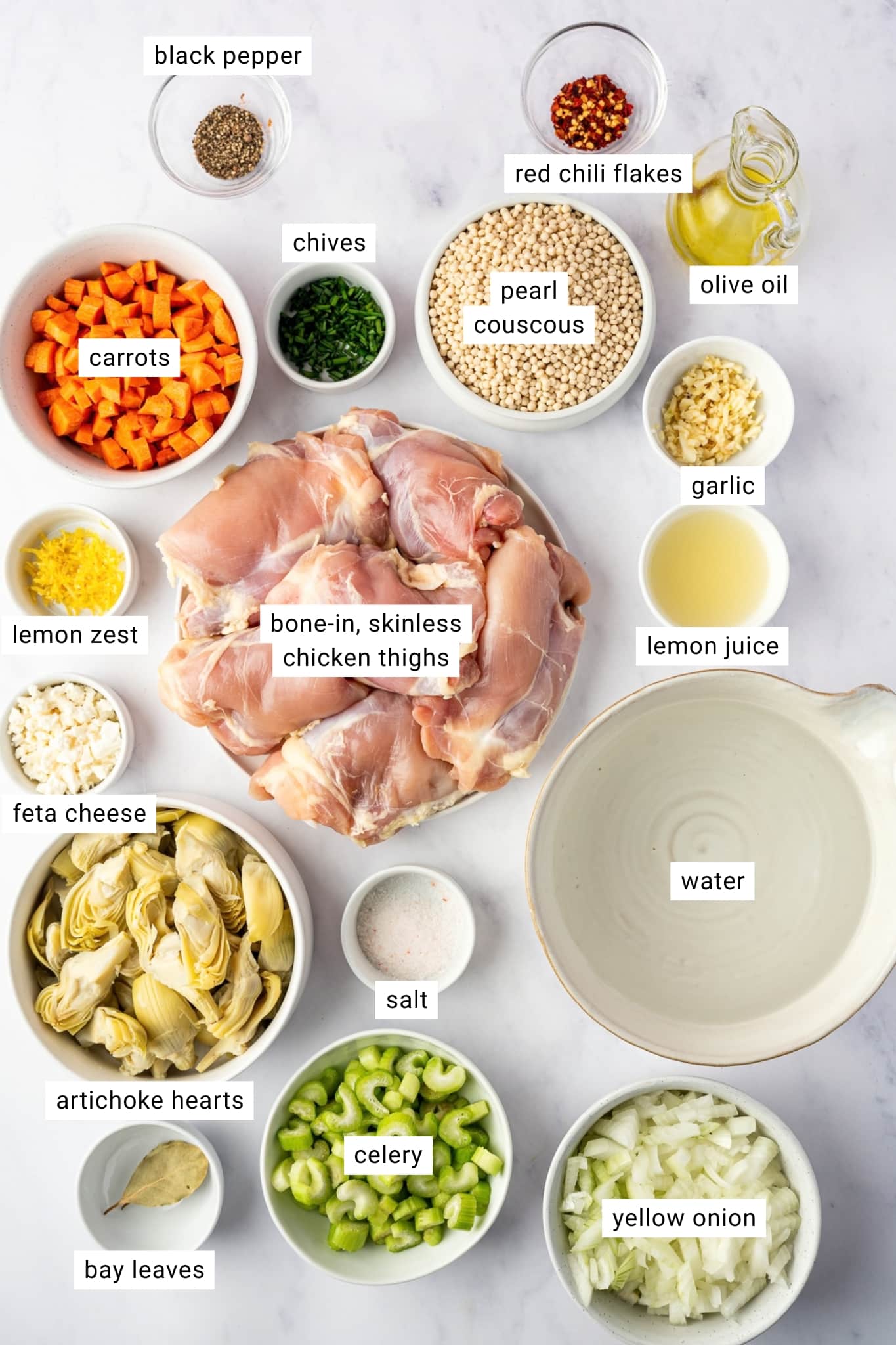 Ingredients for lemon chicken soup arranged in bowls and labeled: bone-in skinless chicken thighs, carrots, celery, yellow onion, garlic, pearl couscous, artichoke hearts, lemon juice, lemon zest, water, olive oil, bay leaves, salt, black pepper, red chili flakes, chives, and feta cheese.