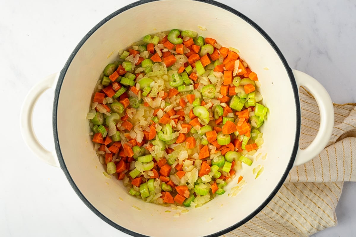 Chopped carrots, celery, and yellow onion softened together in the bottom of a white Dutch oven to create the vegetable base for lemon chicken soup.