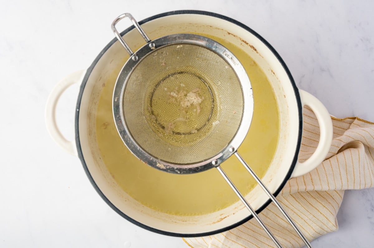 Fine-mesh strainer resting over a white Dutch oven filled with golden homemade chicken broth, catching small pieces of chicken and other solids after the chicken has finished cooking.