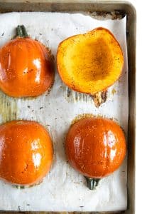 Overhead view of four orange sugar pumpkin halves arranged on a parchment-lined metal baking sheet, with three positioned skin-side up and one showing seasoned orange flesh.