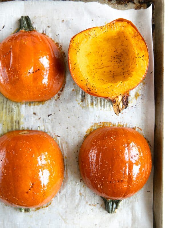Overhead view of four orange sugar pumpkin halves arranged on a parchment-lined metal baking sheet, with three positioned skin-side up and one showing seasoned orange flesh.