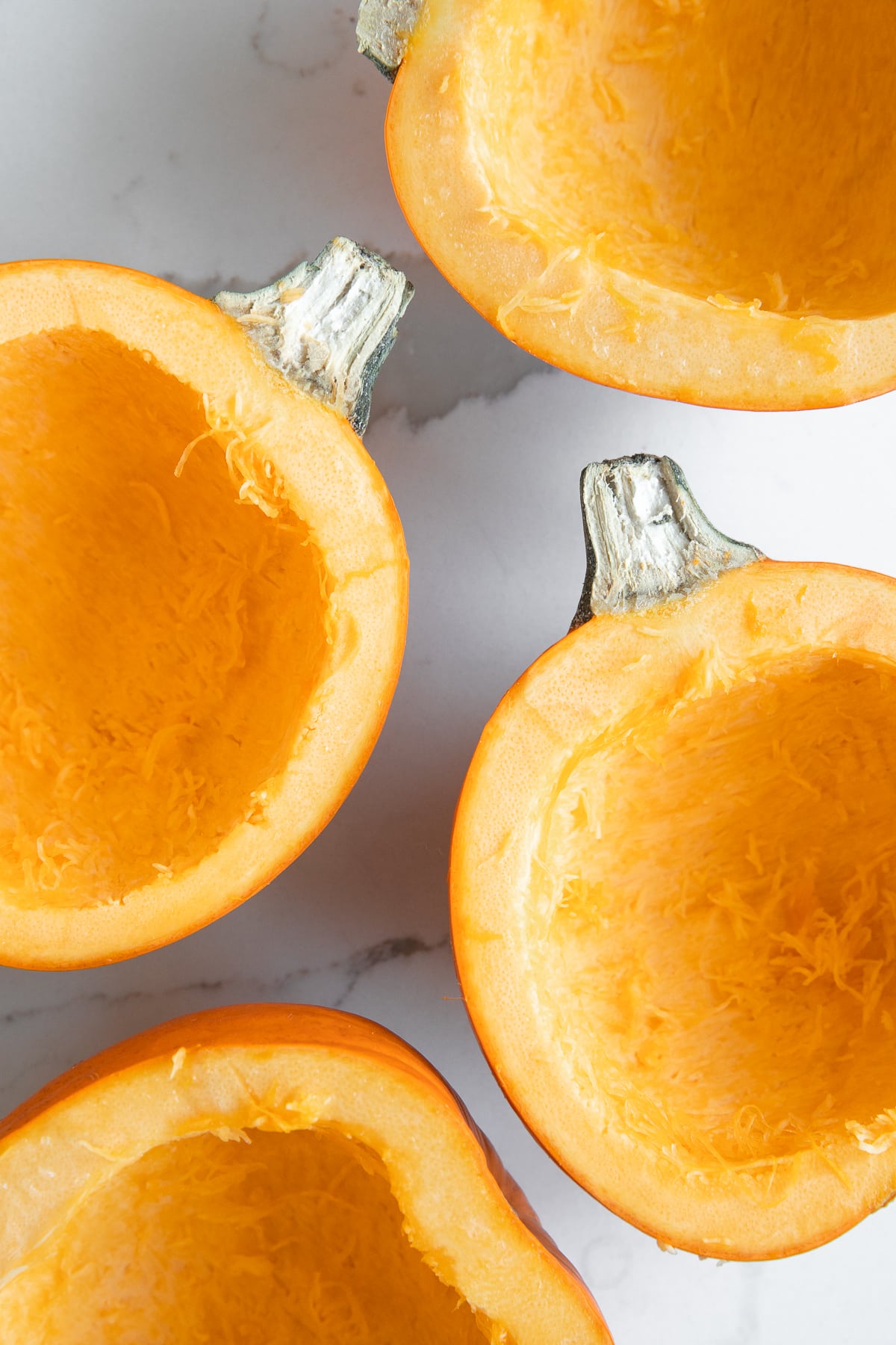 Overhead view of four orange pumpkin halves with the seeds and stringy centers completely scooped out on a white marble countertop.