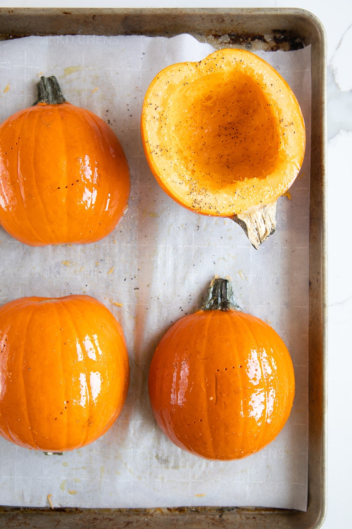 Overhead view of orange sugar pumpkin halves on a parchment-lined metal baking sheet, with the cut sides brushed with oil and seasoned with salt and black pepper.